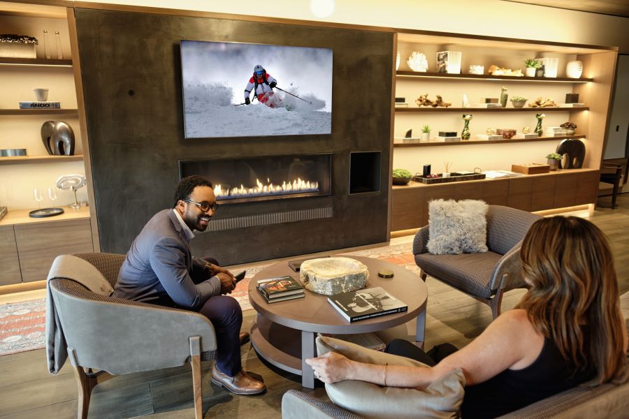 Two people sitting in a living room with the fireplace lit, a TV depicting skiing, and Josh.ai’s control on the wall.