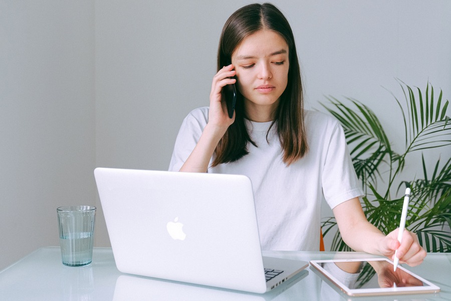 Woman sitting at her desk, a laptop open in front of her, holding her phone to her ear and writing something down.