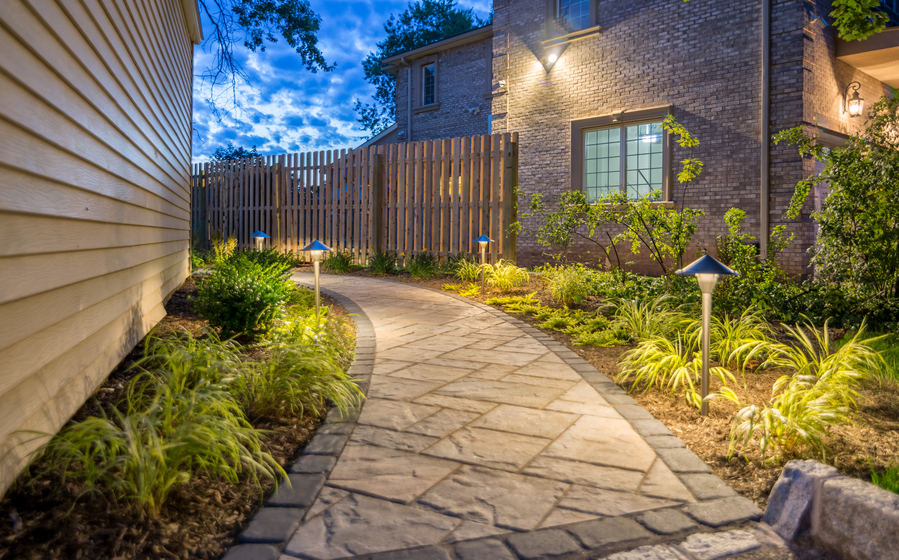 A beautifully lit stone walkway winds past landscaped garden beds and leads to a brick home, creating a warm, inviting outdoor ambiance.