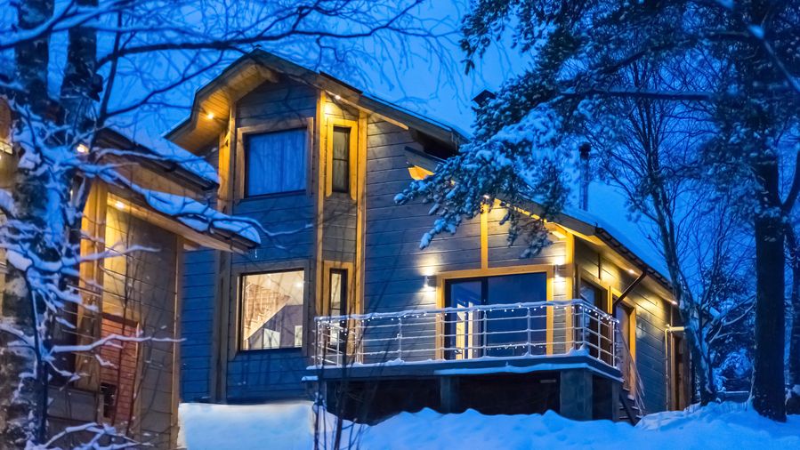 Modern cabin-style home in a snowy landscape at dusk, warmly lit from within and surrounded by trees.