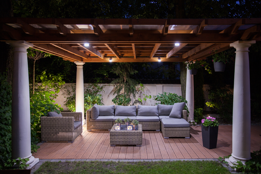 A wicker furniture set is on a wooden deck under a lit-up pergola, with white columns, at night. There are hanging flower pots on the columns.