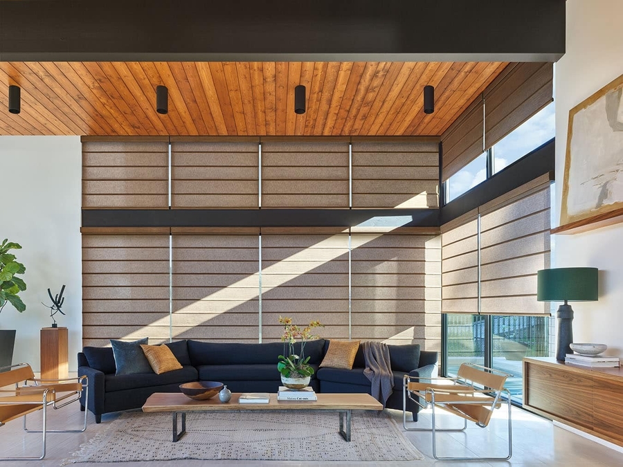 Modern living room with large windows featuring motorized fabric shades, wood-paneled ceiling, neutral furnishings, and abundant natural light.